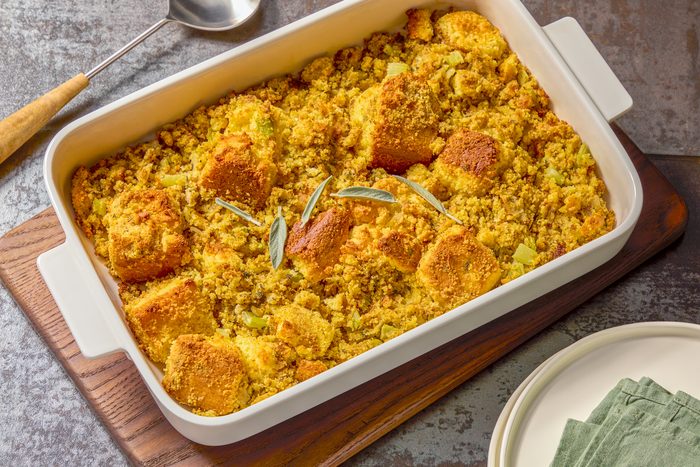 A close-up shot of a white casserole dish filled with a golden brown casserole, The dish is sitting on a dark wooden surface with a light gray background, There is a silver serving spoon sitting on top of the casserole.