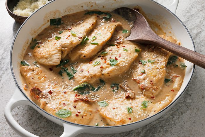 3/4th shot of a white ceramic dish with a black rim, filled with chicken in a creamy sauce, There are also some green leaves and a wooden spoon in the dish, The dish is on a light-colored countertop, The dish is mostly filled with creamy sauce, with the chicken pieces and green leaves scattered throughout, The wooden spoon is lying on top of the chicken, partially submerged in the sauce;