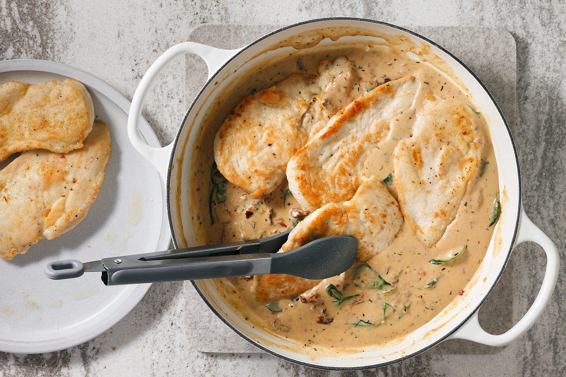 overhead shot of a white ceramic pot with a cream sauce and chicken breast in it, There is a pair of tongs and a plate with chicken breast on the side, The pot is on a grey surface that has a white speckle pattern to it;