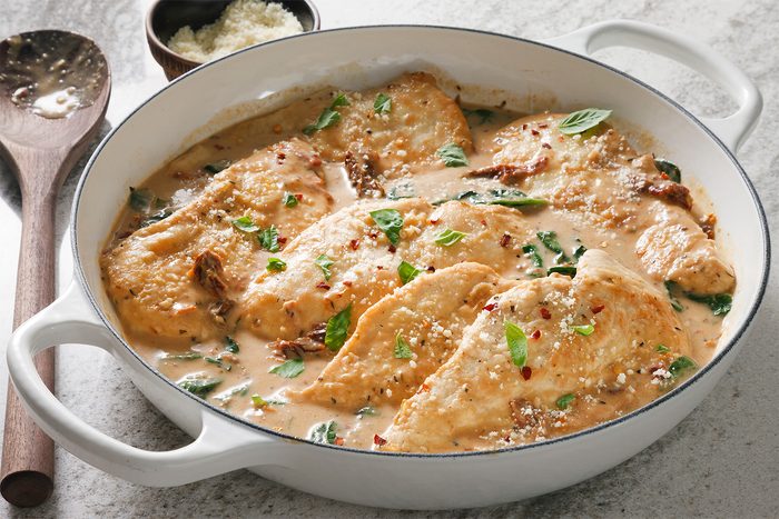 3/4th shot of a white casserole dish with chicken and a creamy sauce, The chicken is cooked and appears juicy, and the sauce is rich and creamy, The dish is topped with a sprinkle of Parmesan cheese and fresh basil leaves, There is a wooden spoon in the bottom left corner of the image, There is a ceramic bowl in the background with parmesan cheese;