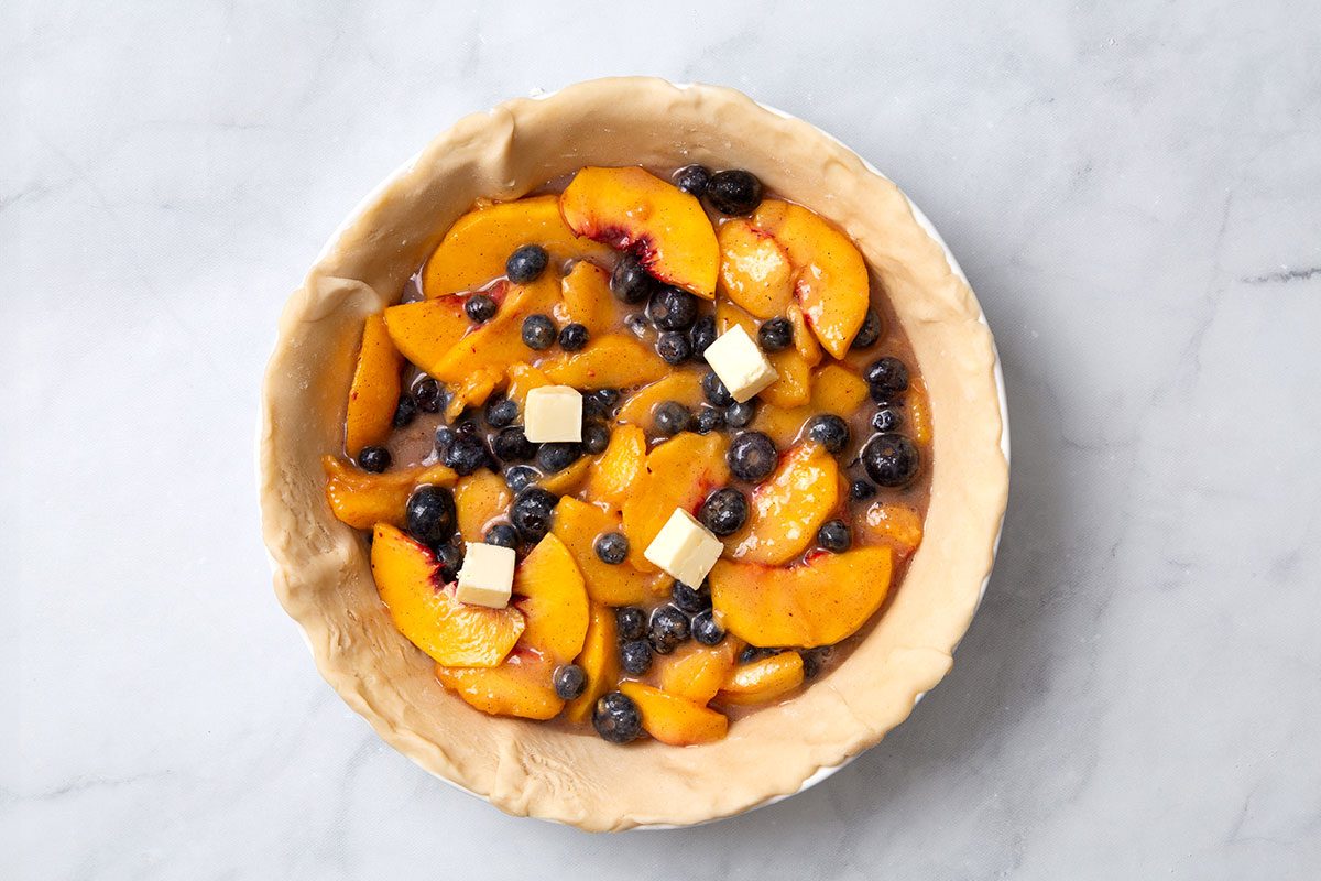 Peach and blueberry filling in a pie pan topped with butter for step three of Peach and Blueberry Pie recipe for Taste of Home
