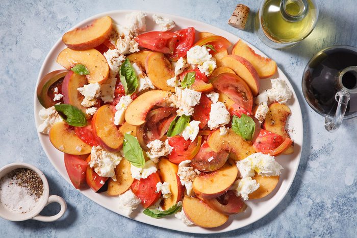A colorful platter of sliced peaches, tomatoes, and chunks of mozzarella cheese is garnished with fresh basil leaves. Nearby are a small bowl of salt and pepper, a bottle of olive oil, and a cork with a shadow on the blue surface.