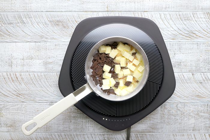 A stovetop with a saucepan containing chopped dark and white chocolate, ready for melting. The surface is wood-textured, and the setup rests on an electric cooktop.
