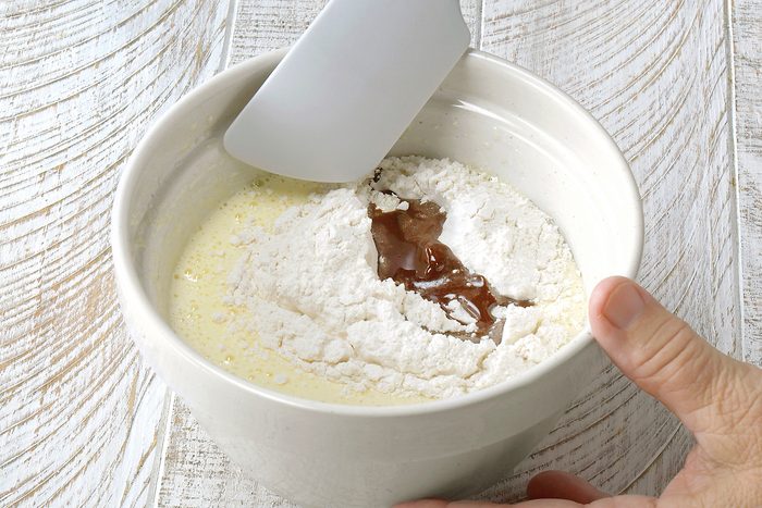 A person holding a white bowl filled with flour and a dark brown liquid, using a spatula to mix the ingredients. The bowl is placed on a light wooden surface.