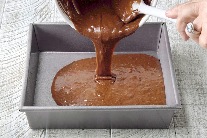 A person pours chocolate cake batter from a white bowl into a greased square baking pan on a wooden surface.