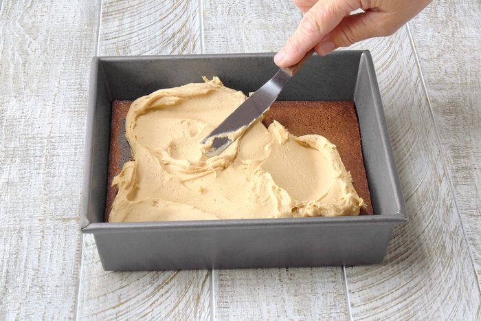 A hand uses a metal spatula to spread creamy frosting over a brown cake in a square metal baking pan on a textured white wooden surface.