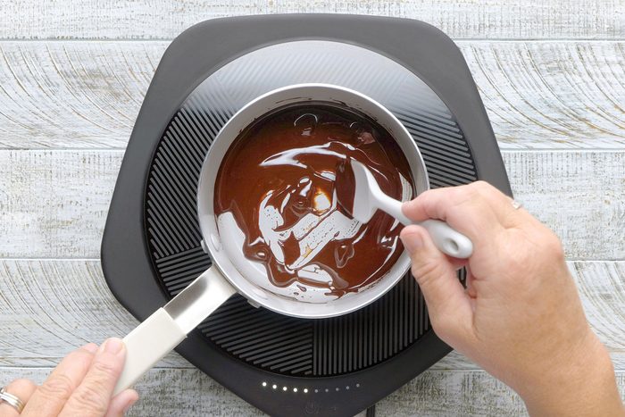 A person stirs melted chocolate in a small saucepan on an induction cooktop. The background is a light, textured wooden surface.
