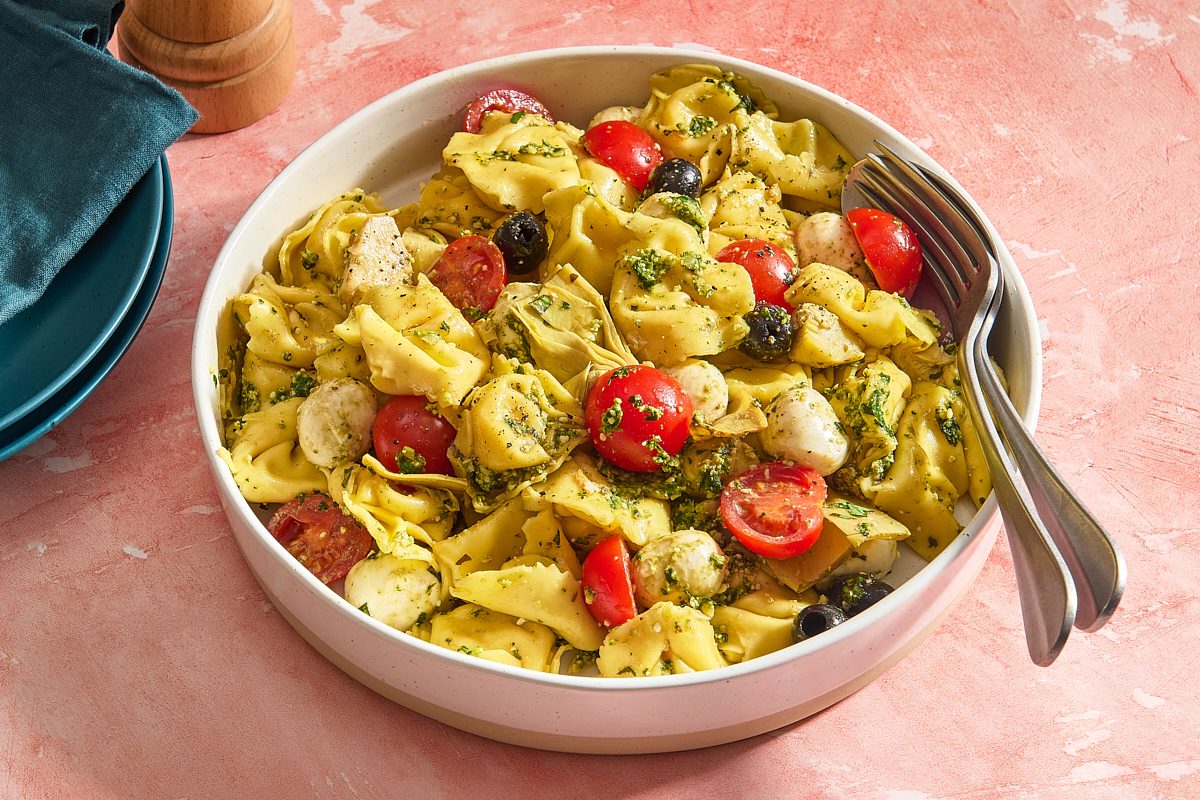 Angled shot of a serving bowl filled with pesto tortellini salad and serving utensils