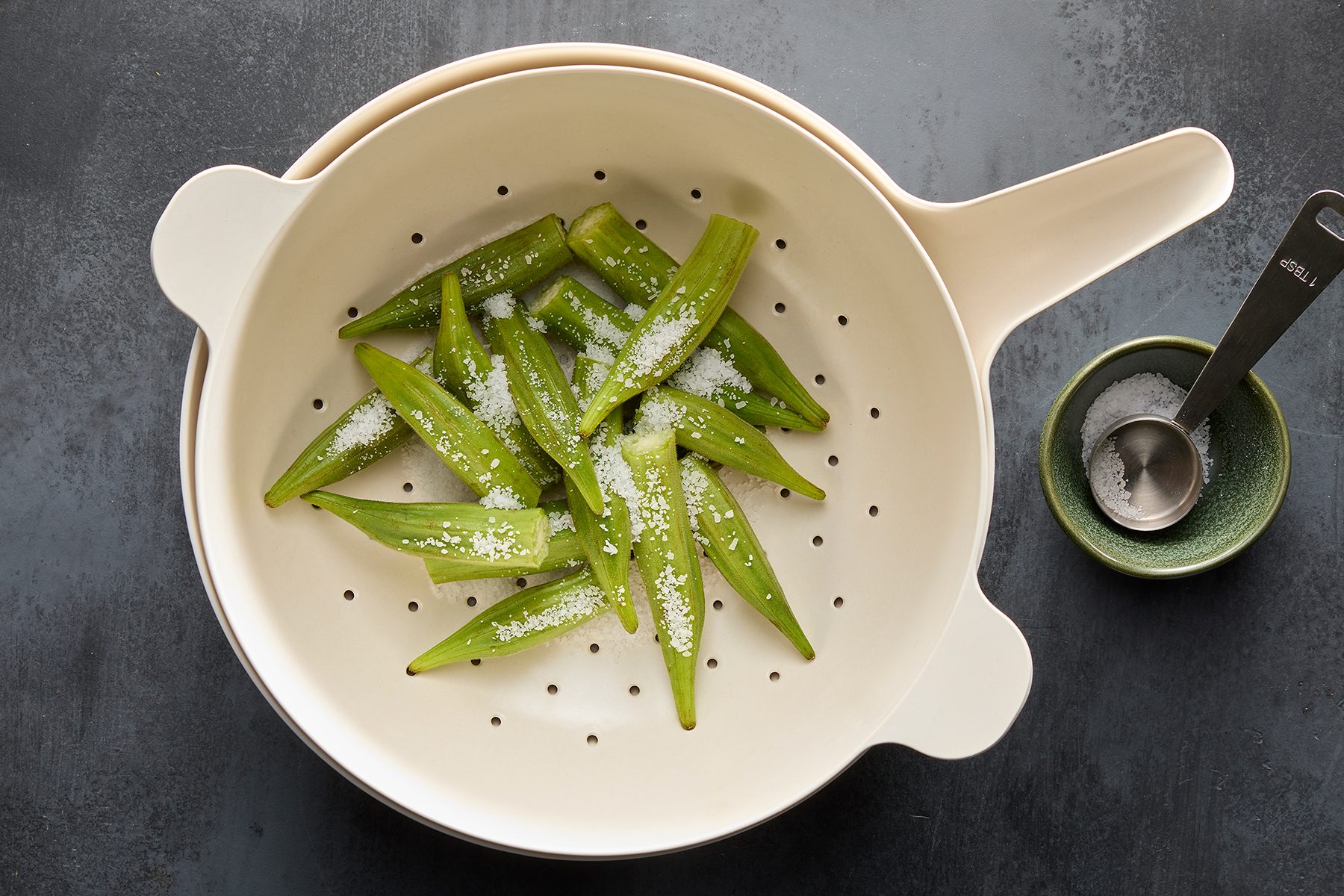 A white colander filled with fresh okra sprinkled with salt is placed on a dark countertop. Next to it, there's a small green bowl with a metal measuring spoon inside.