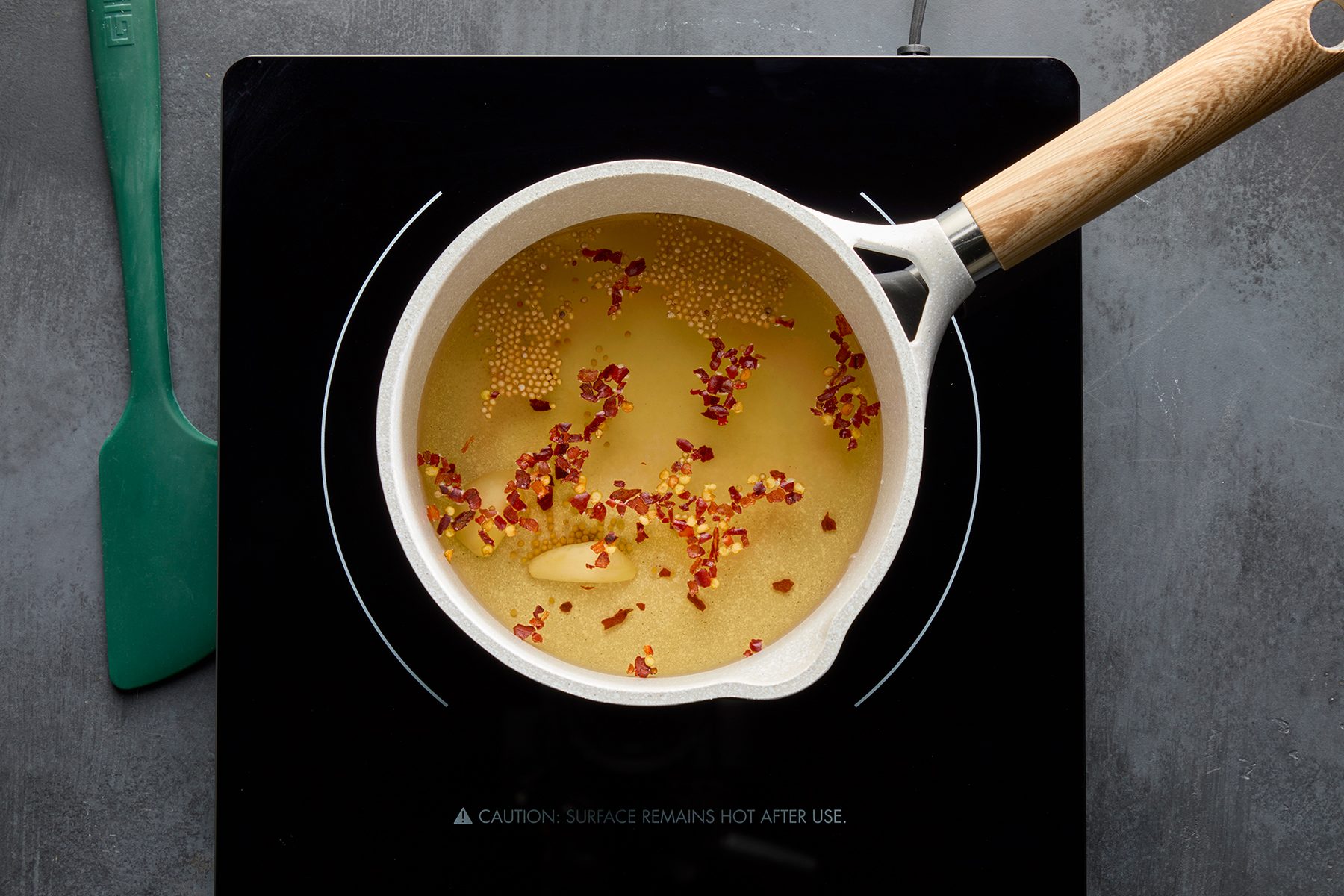 A saucepan with a wooden handle on a black stovetop contains simmering oil with garlic cloves, mustard seeds, and red pepper flakes. A green spatula rests beside the stovetop.