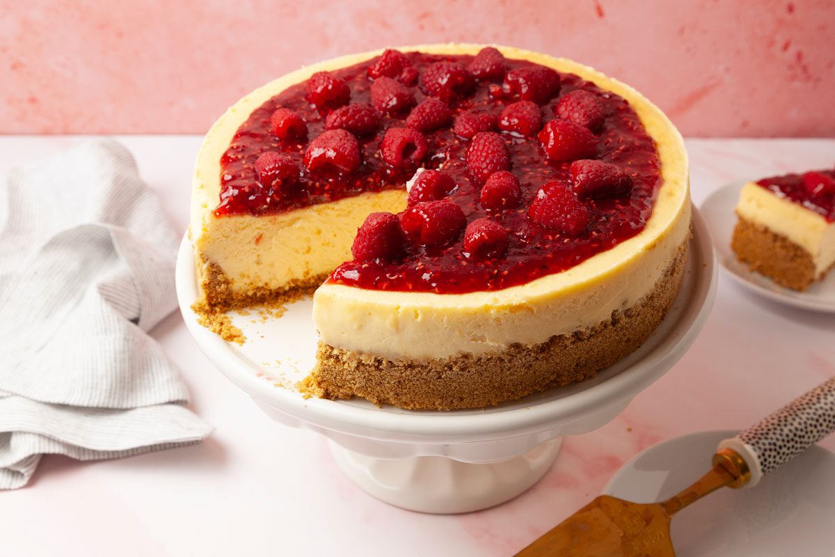 Full yield of Taste of Home Raspberry Cheesecake, pink background, white and black pie server, one slice taken out and in the background