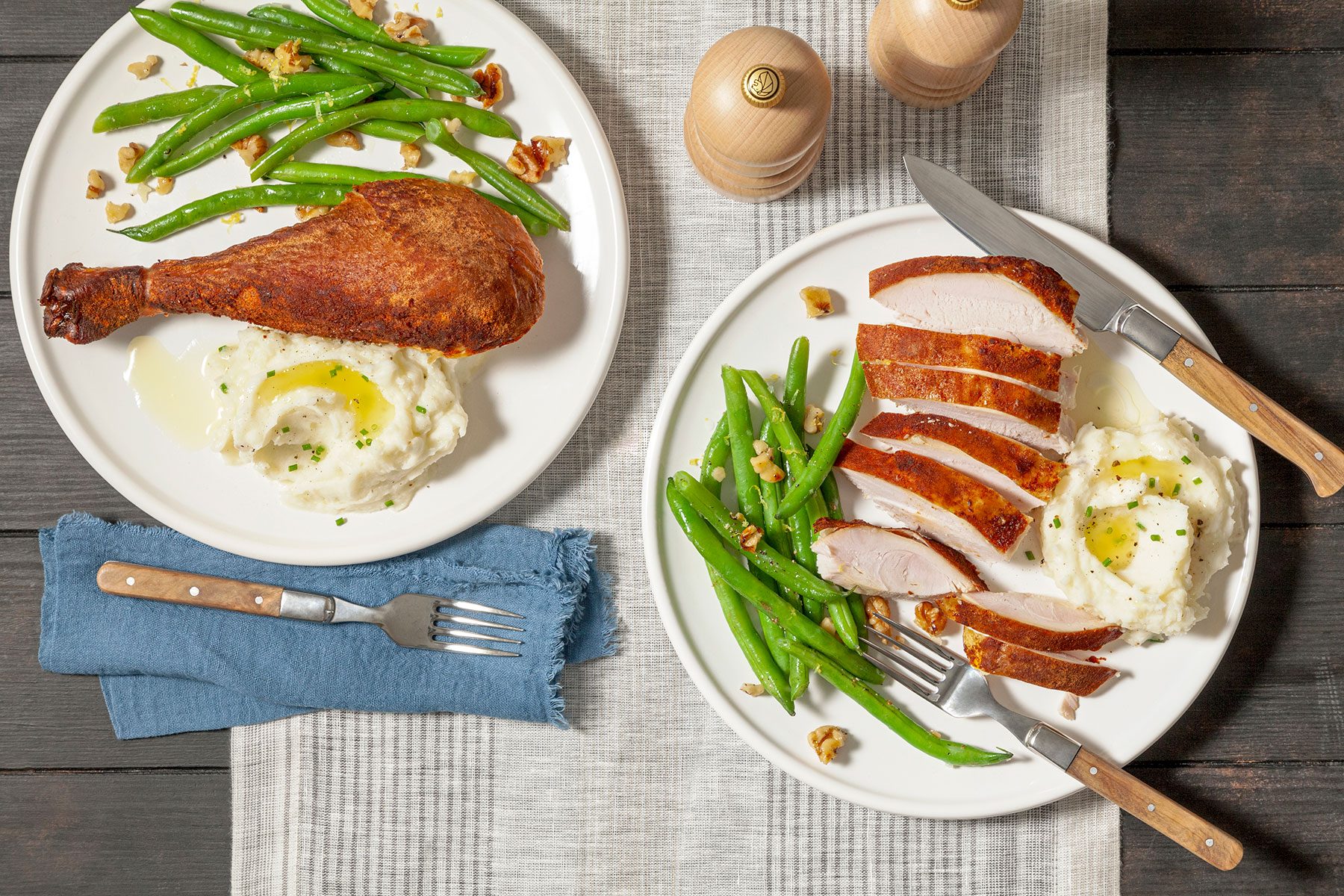 Two plates on a table: one with a roasted turkey leg, mashed potatoes, and green beans; the other with sliced turkey breast, mashed potatoes, and green beans. A fork and knife are by the plates, and two pepper mills are in the background.