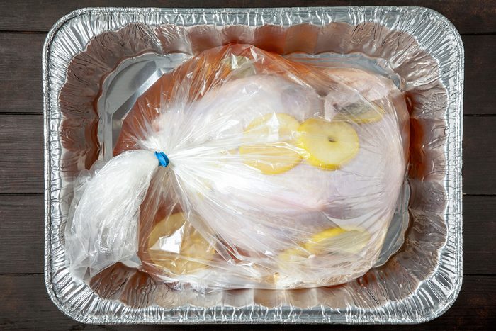 A large raw turkey is placed in a plastic cooking bag with lemon slices. The bag is sealed with a blue tie and rests in a rectangular aluminum roasting pan on a dark wooden surface.