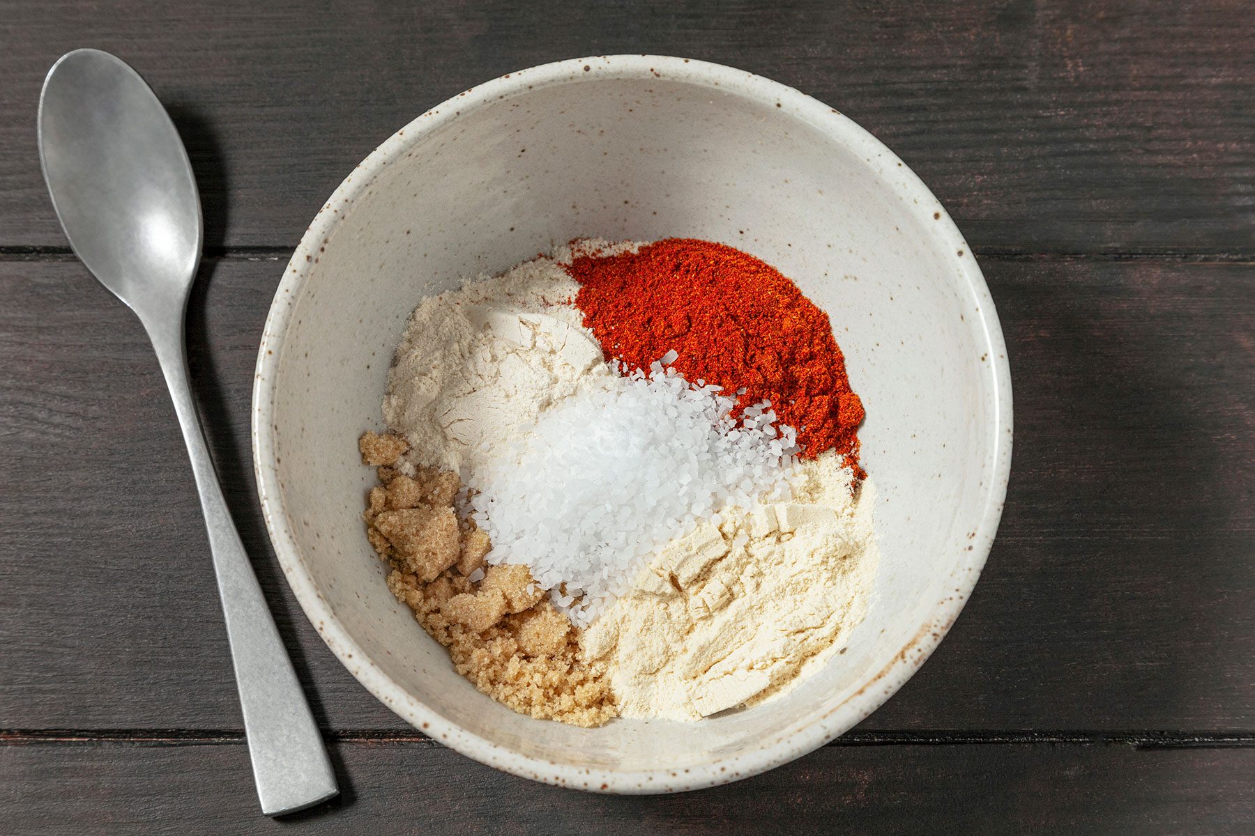 A bowl containing a mixture of spices: salt, brown sugar, flour, and red paprika powder. A silver spoon lies beside the bowl on a dark wooden surface.
