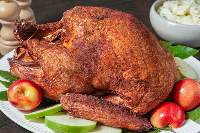 A roasted turkey on a white platter is garnished with green apple slices and red apples. In the background, there's a bowl of mashed potatoes. The turkey's skin is golden brown and crispy.