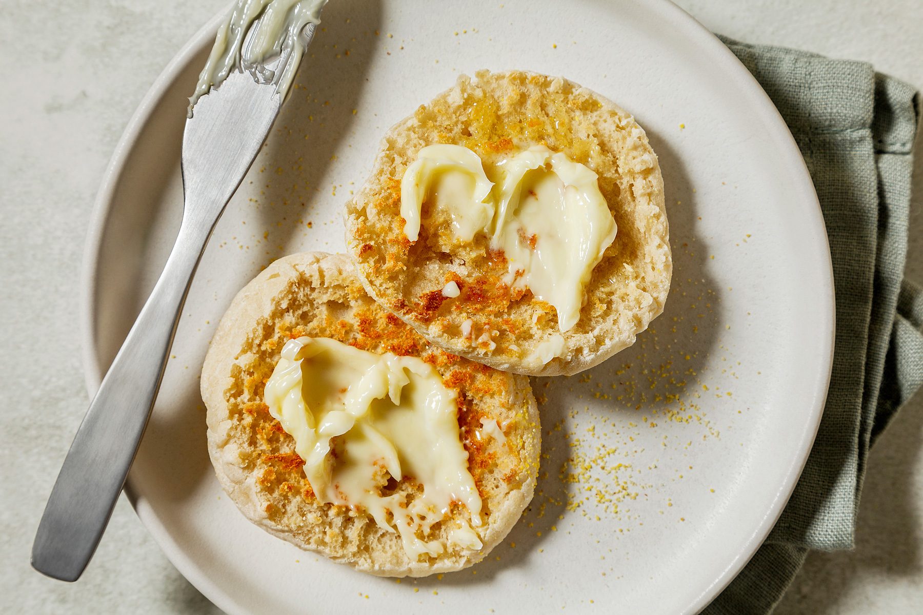 overhead shot of two split English muffins on a plate, The muffins are golden brown and have a slightly crispy exterior, They are topped with melted butter, which is dripping down the sides. A knife is resting on the plate;