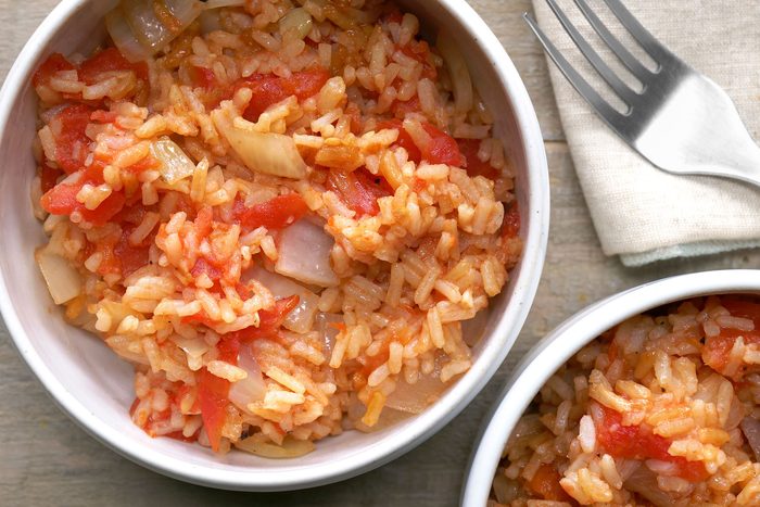 Two bowls of rice pilaf, The rice is a light brown color and is mixed with chopped tomatoes, onions, and herbs, The rice grains are distinct and the vegetables are visible throughout the dish, a silver fork is placed on kitchen napkin