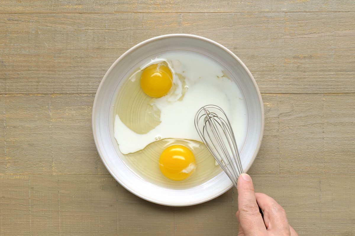 overhead shot of eggs and milk being whisked in a shallow bowl on a wooden surface