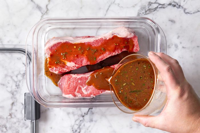 overhead shot of raw steaks in a clear glass container on a marble surface, A hand is pouring a rich, herb-flecked marinade over the meat from a glass measuring cup, The raw steaks are a vibrant pink color;
