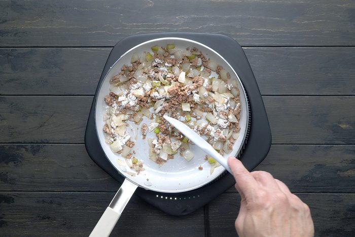 overhead shot of A white skillet contains a mixture of browned ground meat, chopped onions, and chopped celery, A sprinkle of flour is being added to the mixture, and a spatula is being used to stir it in;