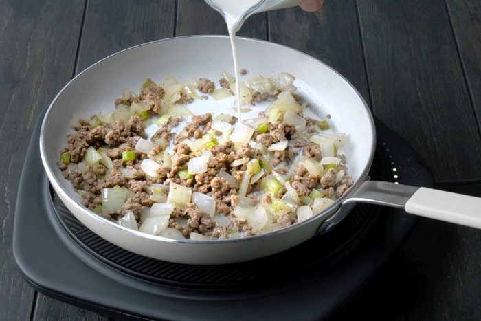 3/4th shot of a white skillet on an electric induction cooktop, The skillet contains a mixture of browned ground beef, chopped onions, and chopped celery, A person's hand is pouring a liquid, likely milk or cream, into the skillet;