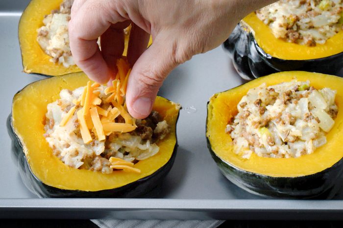 3/4th shot of a baking sheet filled with stuffed acorn squash, A person's hand is sprinkling shredded cheese over one of the stuffed squash halves, The squash shells are a vibrant orange color, and the filling is a mixture of ground meat, rice, and vegetables, The melted cheese is creating a golden brown topping on the squash;