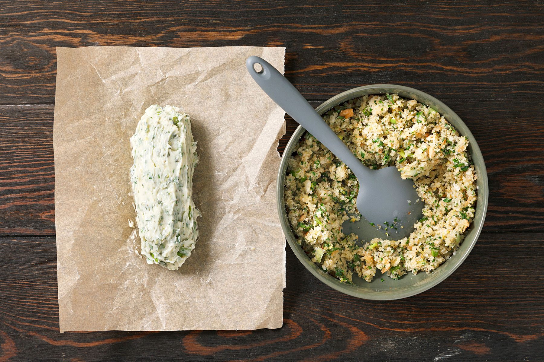 A bowl of couscous mixed with herbs and vegetables sits on a dark wooden table. Next to it, a log of herb butter is placed on a piece of parchment paper with a gray spoon resting inside the bowl.