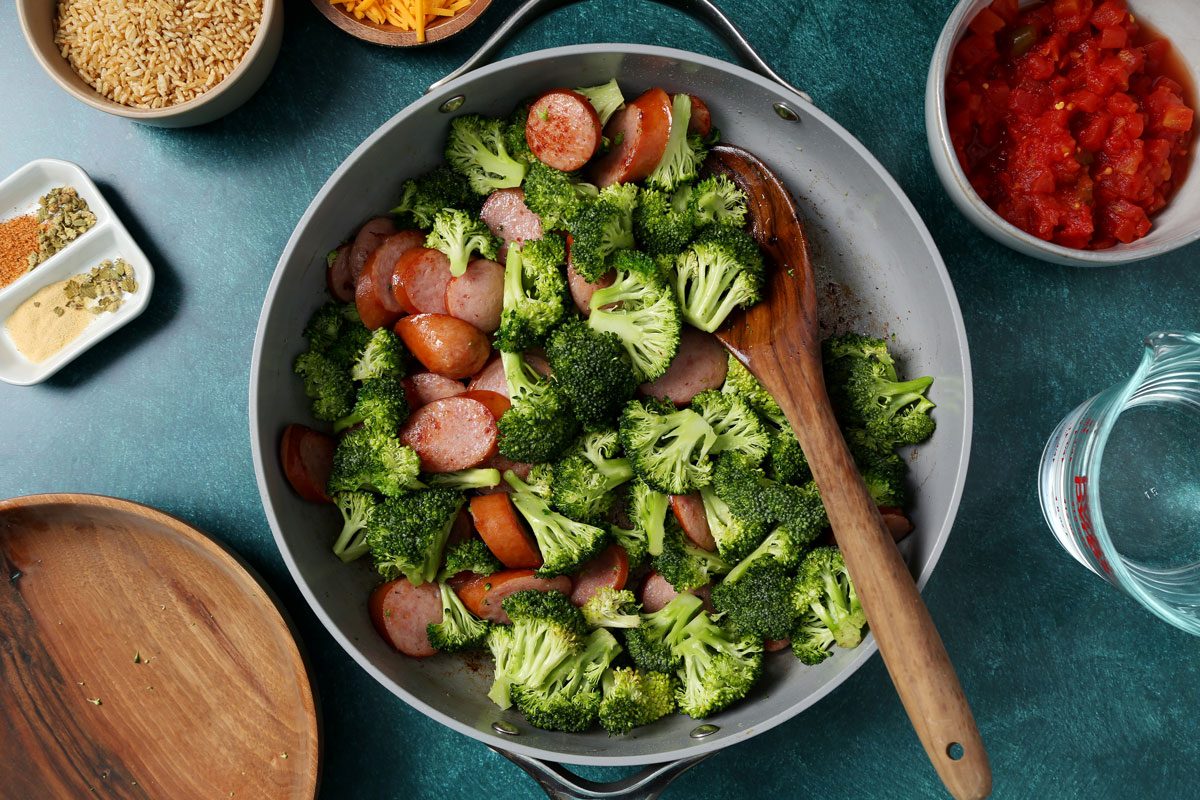 Process of making Taste of Home's Sausage, Rice and Broccoli in a large skillet on a green surface with a green linen.