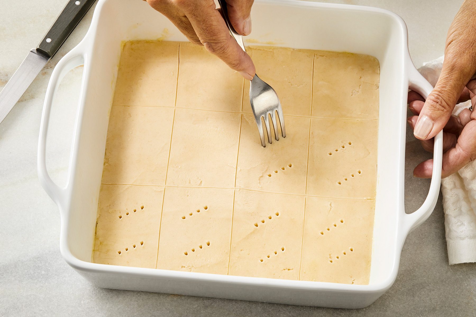 3/4th shot of Using a knife to score batter into 2 in squares; Using a fork to prick a pattern onto the top of each rectangle in a white baking dish;