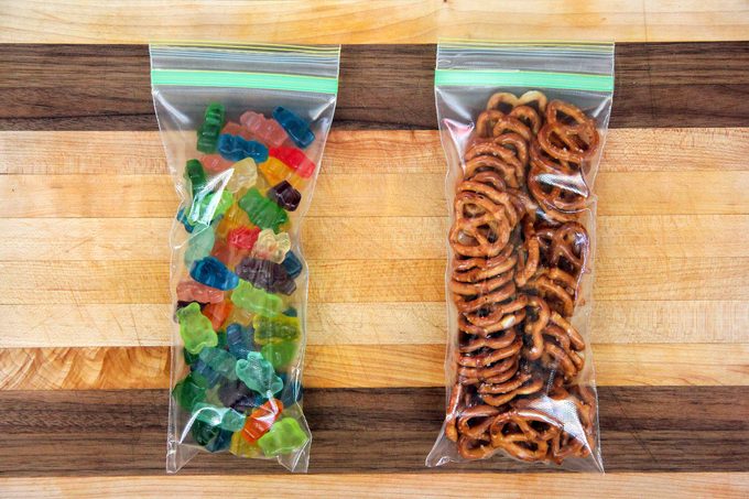 Two clear ziplock bags on a wooden surface: the left bag contains colorful gummy bears, and the right bag holds small pretzel twists.