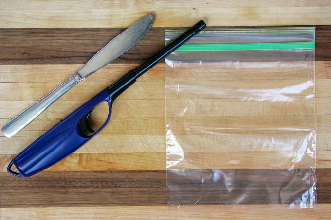 A butter knife, a long lighter, and a clear resealable plastic bag are placed on a wooden cutting board with alternating light and dark wood stripes.