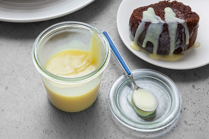 High angle view shot of White Chocolate Sauce; transfer into jar with lid; spoon over cake piece; grey surface;