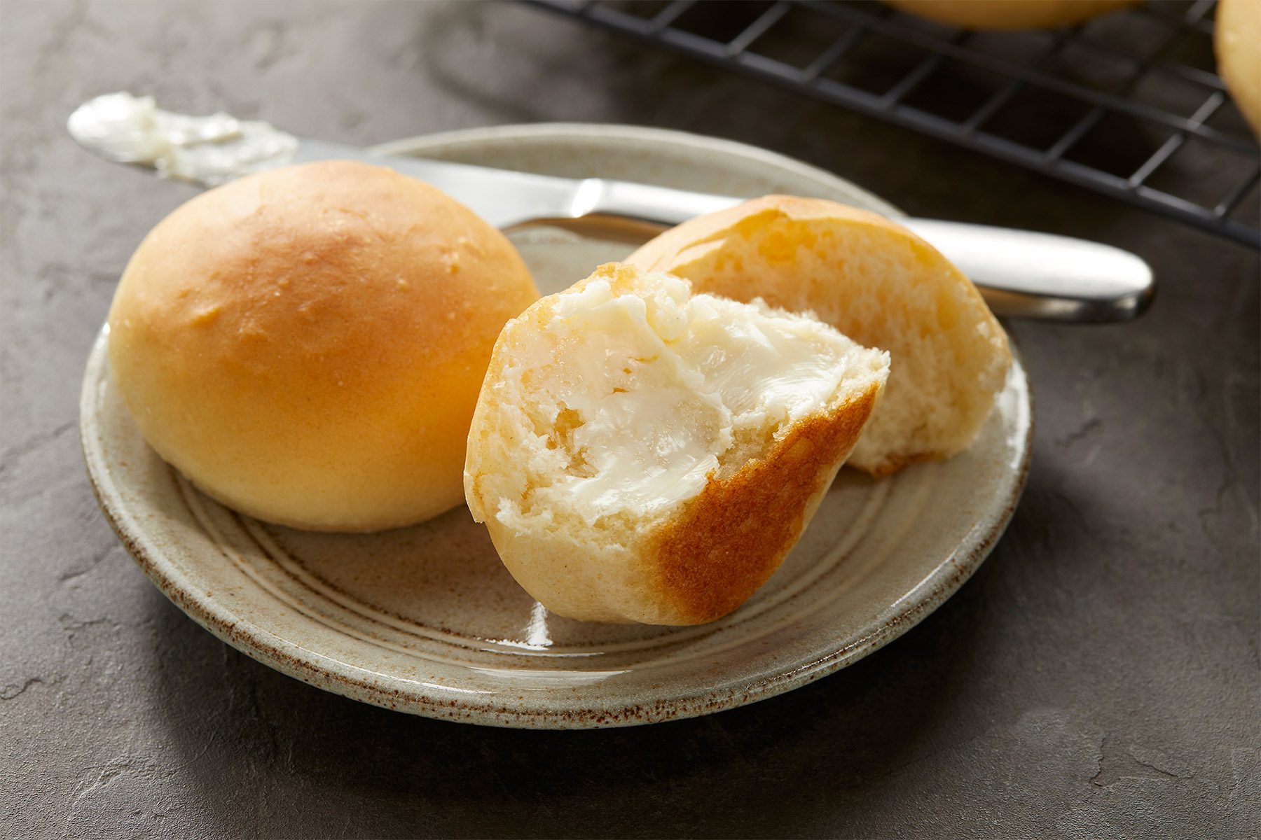 A small plate with two dinner rolls, one whole and one partially sliced open with visible butter spread inside. A butter knife with butter residue lies on the plate. The background shows a cooling rack.