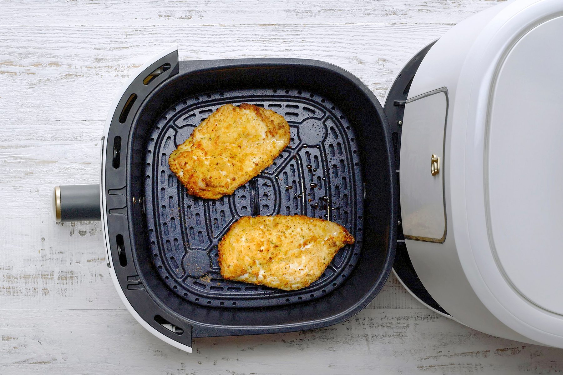 Top view of an air fryer open on a white wooden surface, featuring two cooked, seasoned chicken breasts inside the fryer basket.