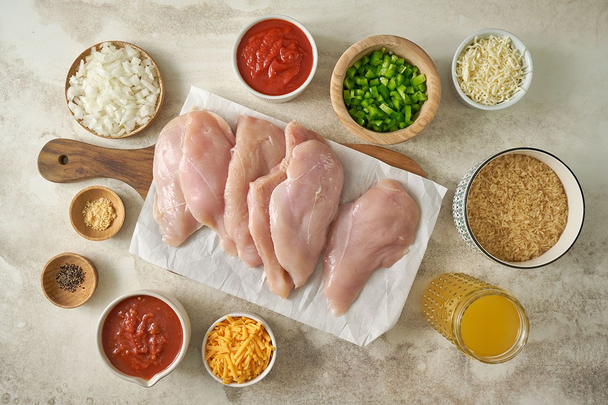 Overhead view of all ingredients for the Taste of Home Arroz con Pollo recipe, including uncooked rice, chicken breast halves, garlic salt, pepper, chicken broth, picante sauce, tomato sauce, chopped onion, green pepper, Monterey Jack cheese, and cheddar cheese, neatly arranged on a countertop.