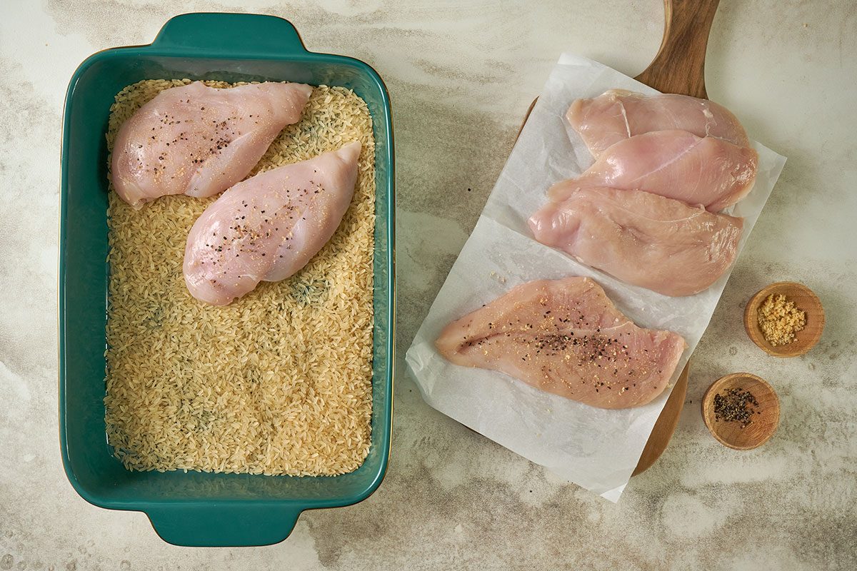 Overhead view of uncooked rice spread in a greased baking dish, with chicken breasts being seasoned with garlic salt and pepper and placed on top of the rice for the Taste of Home Arroz con Pollo recipe.
