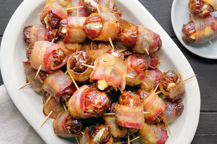 A white plate filled with bacon-wrapped dates, each secured with toothpicks. The dates appear caramelized and arranged neatly, showcasing the crispy bacon exterior. A smaller plate with more dates is partially visible in the background.