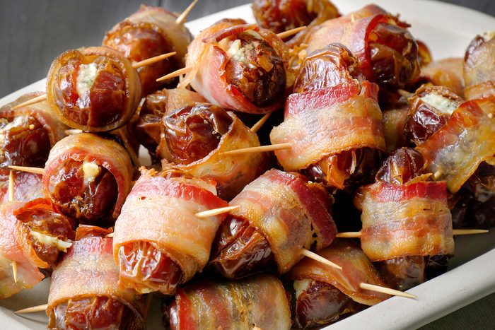 A platter filled with bacon-wrapped dates, each secured with toothpicks. The dates are glistening and the bacon appears crispy. The dish showcases a savory, bite-sized appetizer.
