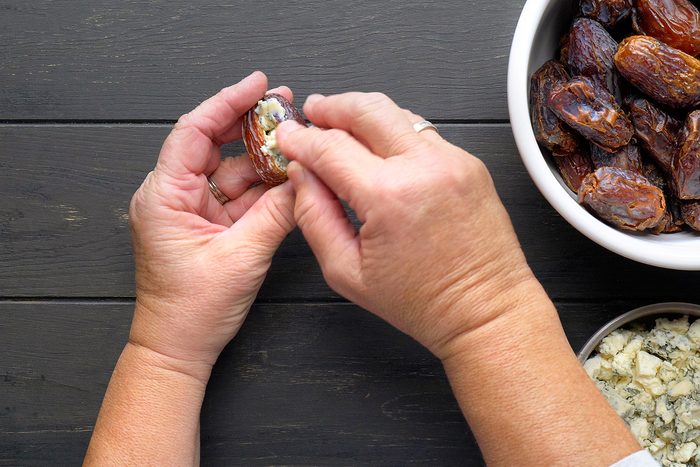 Two hands are stuffing a date with blue cheese. A bowl of dates is on the right, and a bowl of crumbled blue cheese is at the bottom. The background is a dark wooden surface.