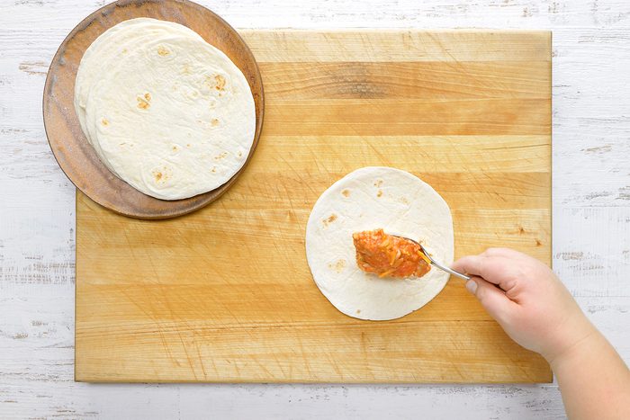 A person's hand spreads a spoonful of sauce on a tortilla. Several tortillas are stacked on a wooden plate to the left. The scene is set on a light-colored wooden surface.