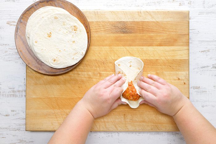 Hands folding a tortilla with filling on a wooden cutting board. A stack of plain tortillas sits on a wooden plate nearby.