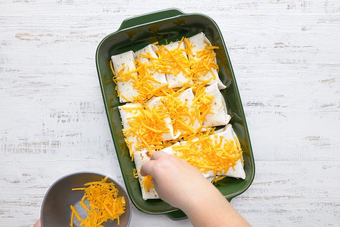 A hand is sprinkling shredded cheddar cheese over six burritos in a green baking dish. The background is a white wooden surface, and there is a small bowl with more cheese in the lower corner.