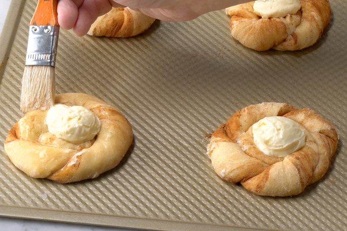 A hand is applying butter with a brush to circular pastries filled with creamy cheese, arranged on a textured baking sheet.