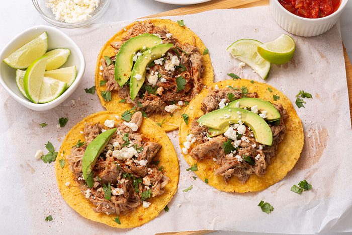 Tostada shells topped with chipotle shredded pork, chopped cilantro, crumbled queso fresco and avocado slices on a wooden cutting board for Taste