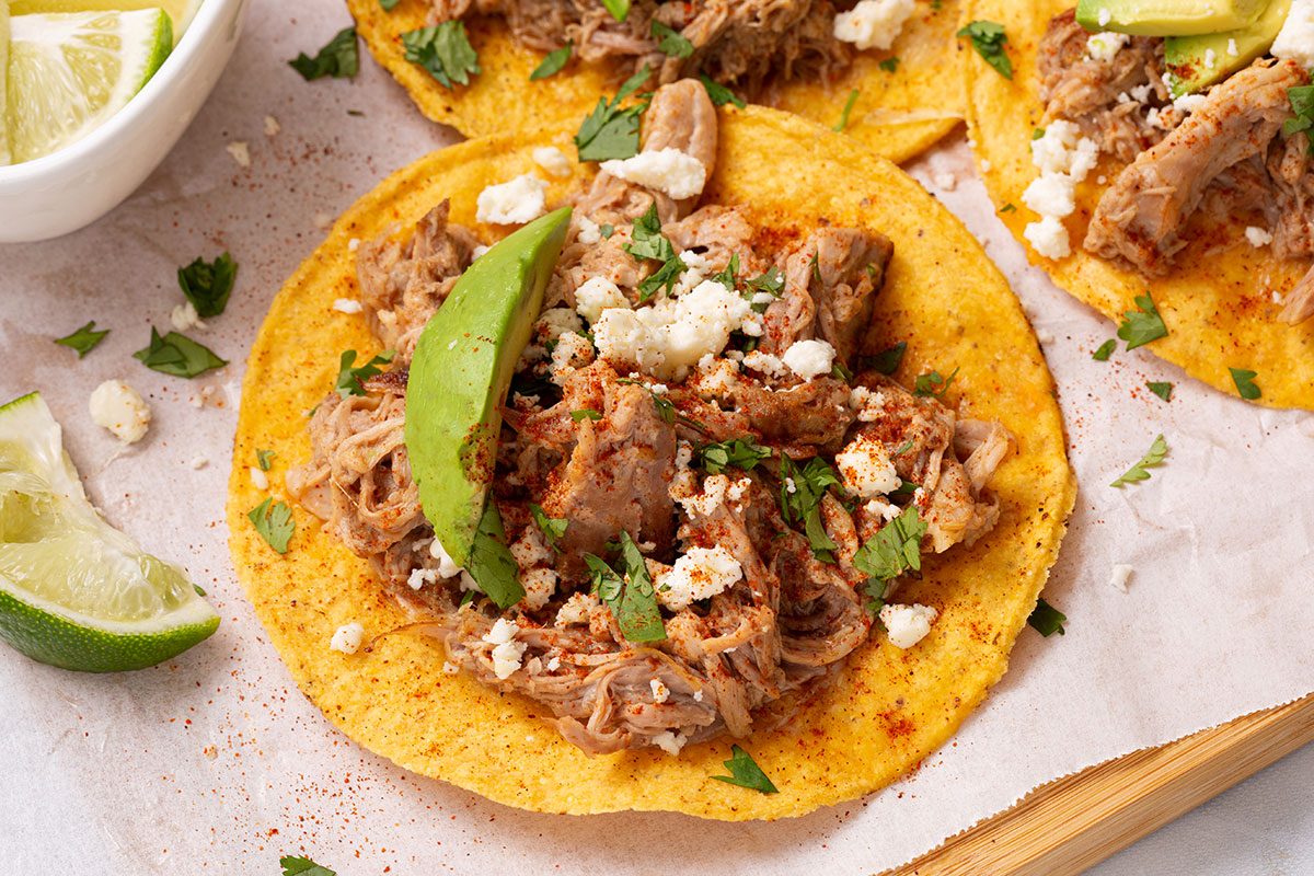 Tostada shell topped with chipotle shredded pork, chopped cilantro, crumbled queso fresco and avocado slices on a wooden cutting board for Taste of Home