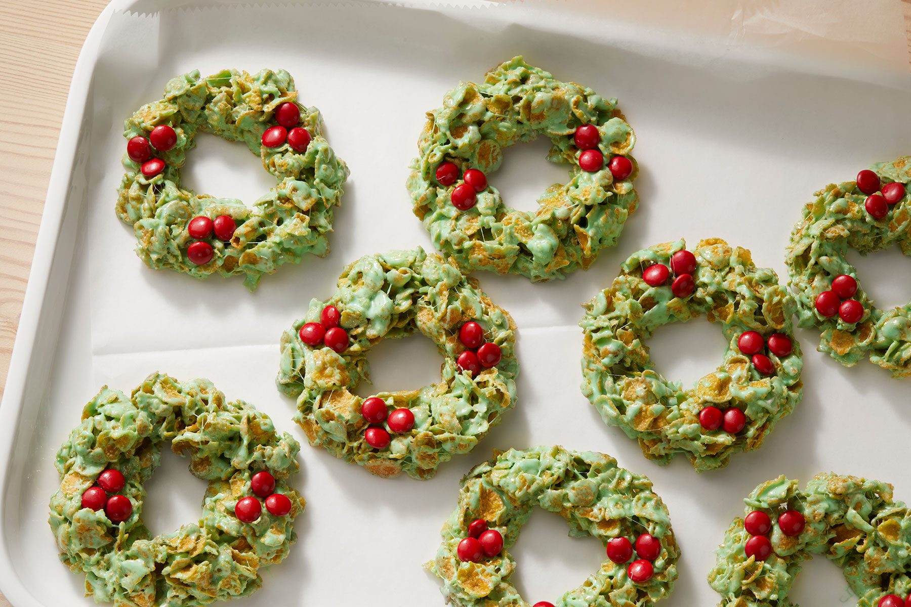 Festive green wreath cookies made from cornflakes are arranged on a tray. Each cookie is decorated with small red candy "berries," resembling holly wreaths.