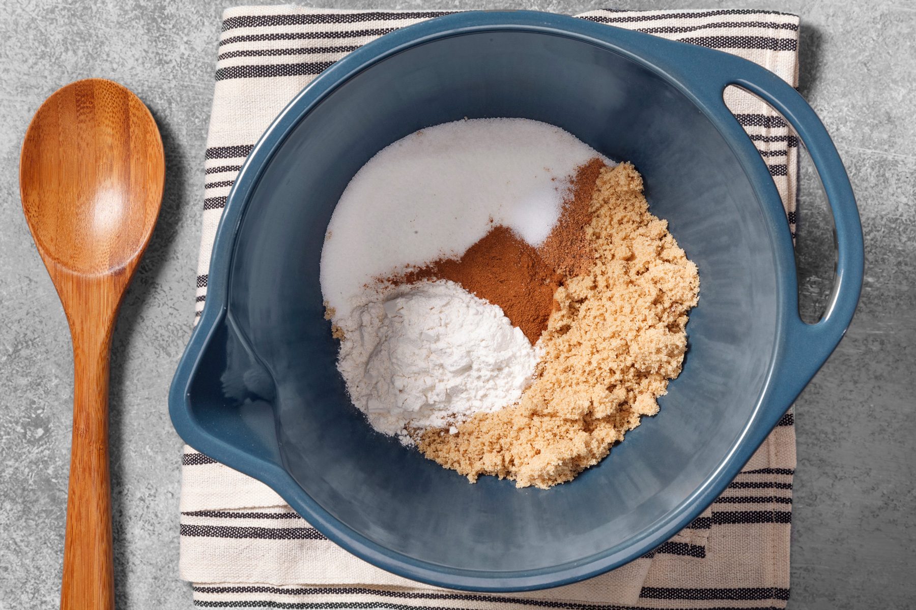 overhead shot of a blue mixing bowl with dry ingredients for baking; there is white sugar, brown sugar, flour, and cinnamon in the bowl; the bowl is on top of a white and black striped kitchen towel; a wooden spoon is next to the bowl on the left side of the image; the background is a light gray textured surface