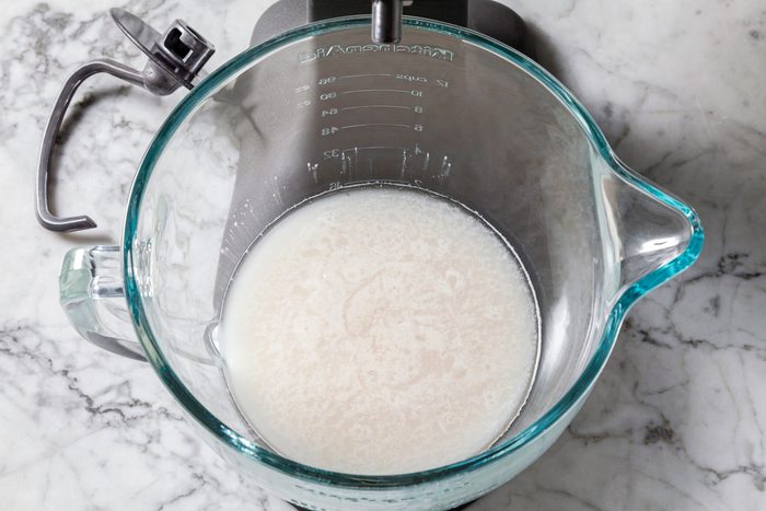 Overhead shot of a stand mixer bowl with hook attachment, warm water and yeast proofing on a marble surface for 5 minutes.