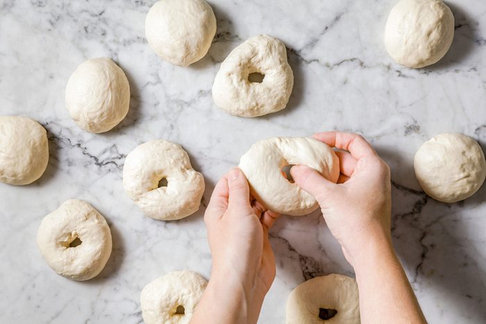 Overhead shot of pressing a thumb through the center of each dough ball, stretching and shaping into an even ring with a 1-inch hole on a marble surface.