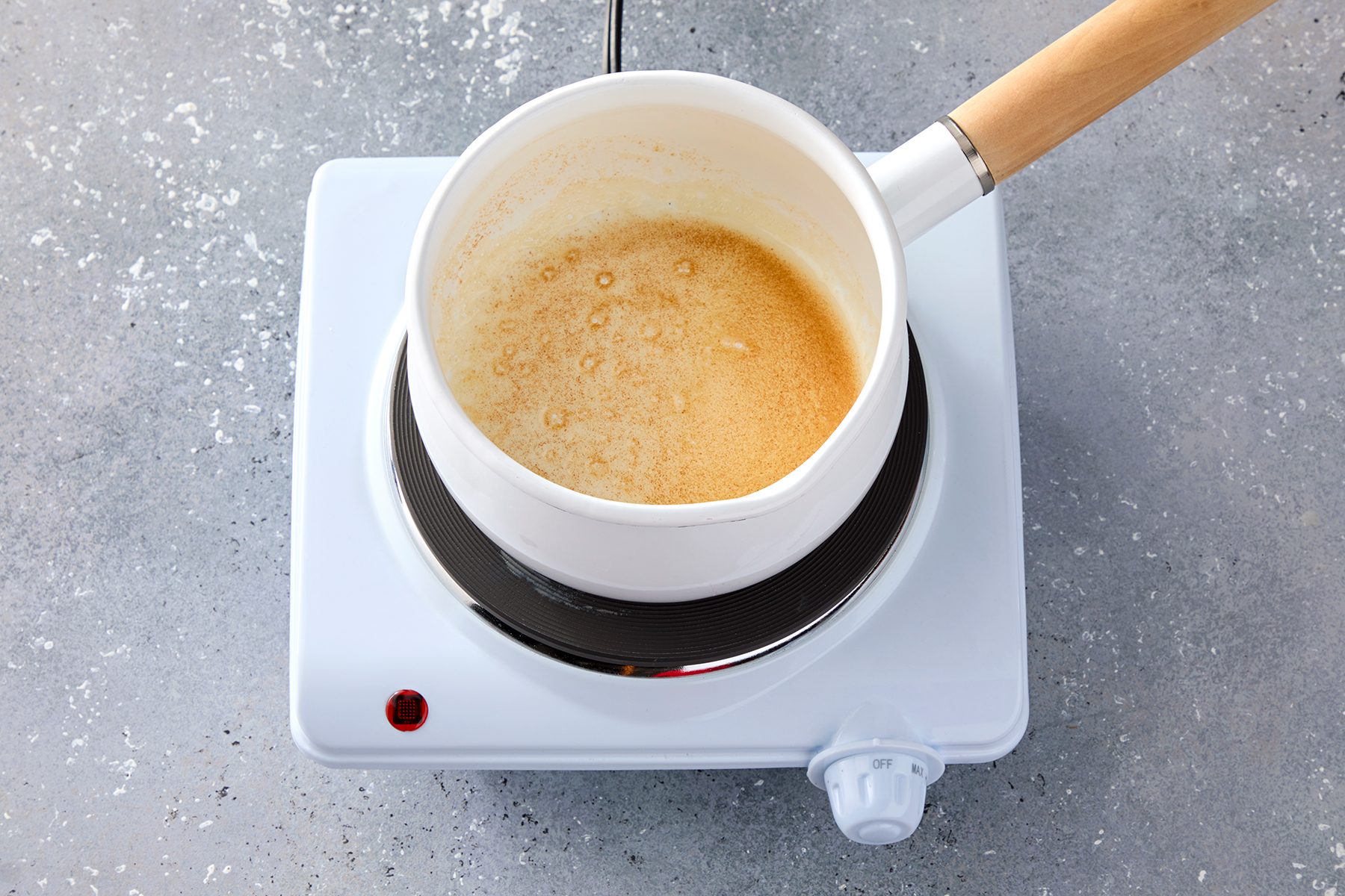 A white saucepan with a wooden handle is placed on a white electric hot plate, containing a bubbling caramel mixture. The background is a speckled gray countertop.